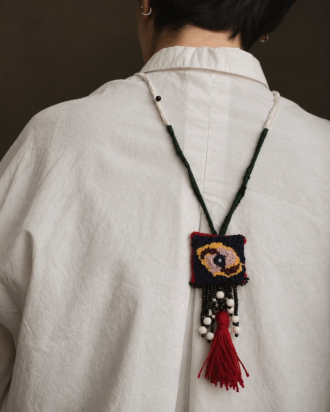 Embroidered eye pendant with beads and a red tassel, worn on the back of a white shirt