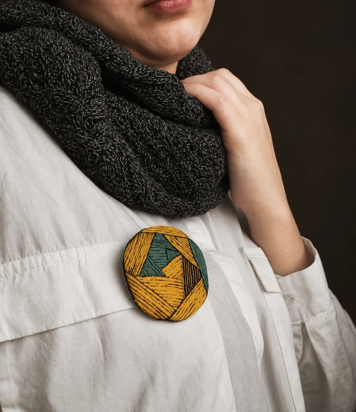 Round embroidered brooch in mustard yellow and green, worn on a white shirt under a grey scarf