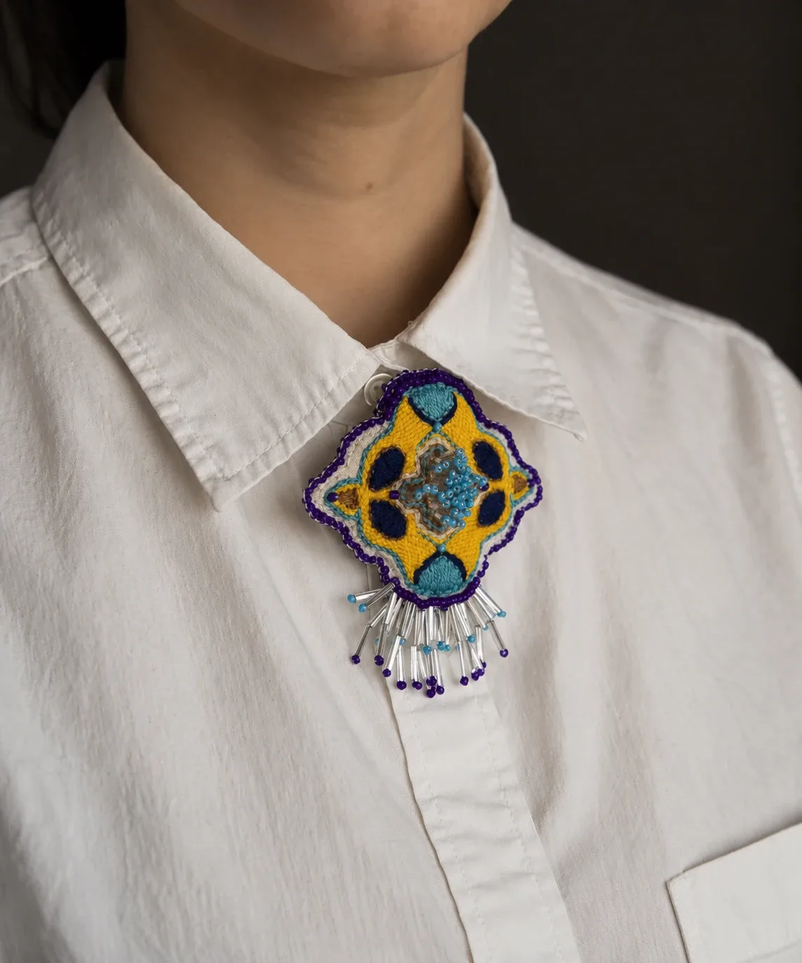 Embroidered brooch in yellow, navy and turquoise with a purple beaded edge and glass bead fringe, worn at the collar of a white shirt